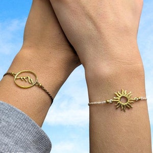 Könnte beinhalten: Zwei passende Freundschaftsarmbänder mit einem goldenen Berg- und Sonnenanhänger an einem weißen und blauen Band. Die Armbänder werden an den Handgelenken von zwei Personen getragen, die ihre Hände gegen einen blauen Himmel erhoben haben.