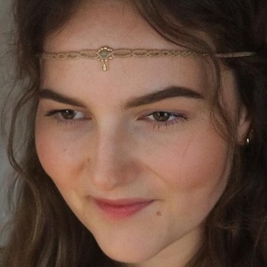 May include: A woman with long brown hair wearing a gold chain headband with a small pendant. The headband is made of thin rope and has a small, round charm in the centre. The woman is looking down and smiling.