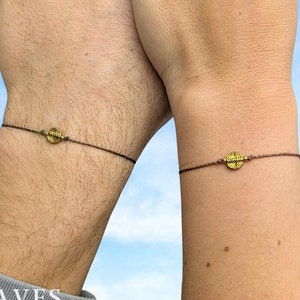 May include: Two matching bracelets with brown cord and a small gold charm. The charms are circular with a design on them.