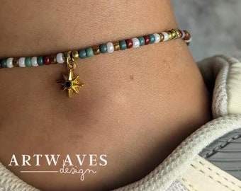 May include: An anklet featuring a repeating pattern of small white, sage green, dark red, and metallic gold beads. A gold-toned star-shaped charm with a small dark centre is suspended from the beaded strand. The text ARTWAVES design is visible.