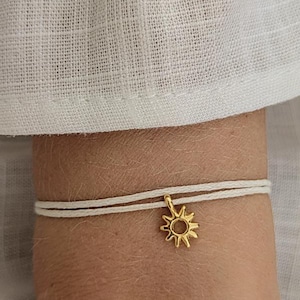 Könnte beinhalten: Ein goldfarbener Sonnenanhänger an einem zweisträngigen weißen Kordelarmband. Der Sonnenanhänger hat eine kreisförmige Mitte mit strahlenförmigen Spitzen. Das Armband ist vor einem weißen Stoffhintergrund mit dem Text ARTWAVES design abgebildet.