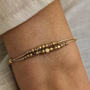 May include: A double-strand bracelet featuring beige cord and a series of metallic gold-toned beads in various shapes, including round and faceted geometric forms, interspersed with small textured grey beads throughout the design.