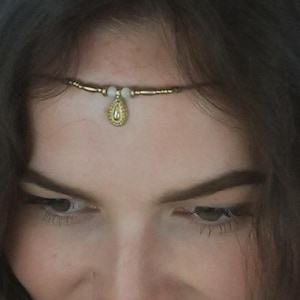 Pode incluir: Uma faixa de cabeça com contas douradas e brancas com um pingente em forma de lágrima. A faixa de cabeça é usada na testa de uma pessoa.