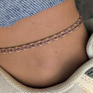 以下が含まれることがあります： 足首に、茶色、ベージュ、グレーの糸で編まれた細いアンクレットをつけた人の足首のクローズアップ。アンクレットは足首で結ばれています。