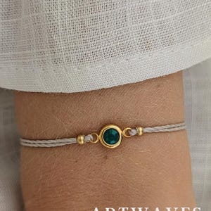 Può includere: Un braccialetto delicato con una montatura circolare dorata centrale che racchiude una singola pietra preziosa rotonda verde scuro. Il ciondolo è affiancato da due piccole perline dorate su un sottile cordino grigio chiaro.