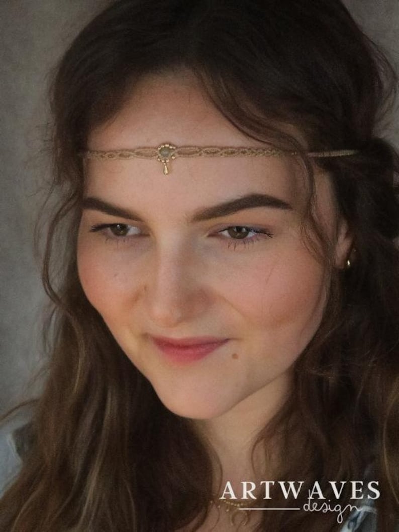 May include: A woman with long brown hair wearing a gold chain headband with a small pendant. The headband is made of thin rope and has a small, round charm in the centre. The woman is looking down and smiling.