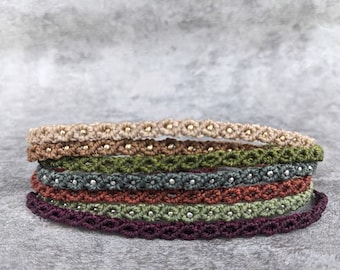 Könnte beinhalten: Ein Stapel aus sechs dünnen Makramee-Armbändern in verschiedenen Farben wie Beige, Braun, Olivgrün, Dunkelgrau, Rostrot und Dunkelviolett. Jedes Armband besteht aus einem geflochtenen Kordelmuster mit kleinen Metallperlen in der Mitte jedes Knotens.