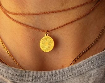 Può includere: Un ciondolo circolare color oro con un design inciso di sole e luna, sospeso a una collana a doppio cordino marrone. Il ciondolo è indossato insieme a una catenina color oro. Il testo ARTWAVES design è visibile nella parte inferiore.