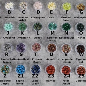 May include: A collection of 25 small containers, each filled with different coloured beads. The containers are labelled with letters A through Z and Z1 through Z5. Each container has a label with the name of the bead type in German.
