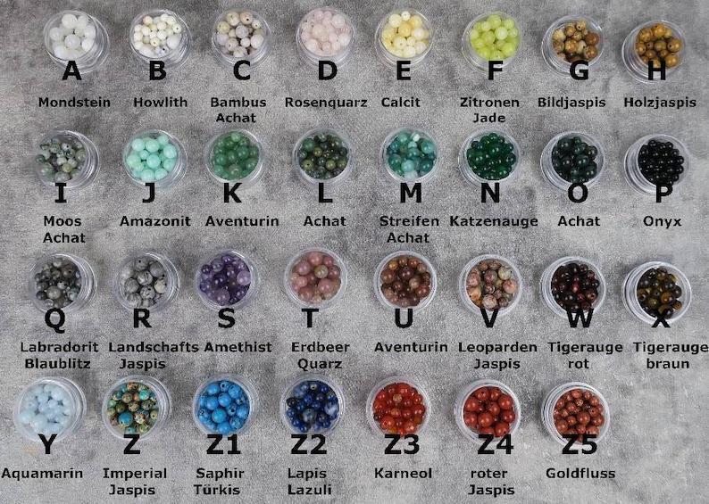 May include: A collection of 25 small containers, each filled with different coloured beads. The containers are labelled with letters A through Z and Z1 through Z5. Each container has a label with the name of the bead type in German.