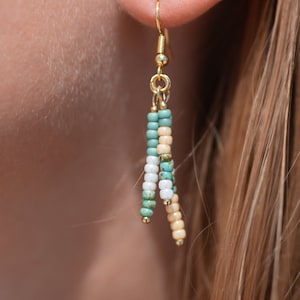 Può includere: Orecchini con perline dorate con uno schema di colori turchese, bianco e crema. Gli orecchini sono appesi all'orecchio di una persona.