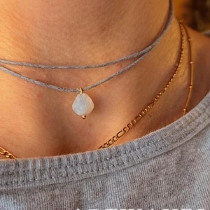 Peut inclure: Un ensemble de colliers superposés composé d'un ras-du-cou en cordon gris fin avec un petit pendentif en pierre translucide en forme de goutte et des détails dorés, accompagné d'une chaîne en métal doré au design à maillons classiques.