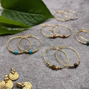 Può includere: Un set di orecchini a cerchio dorati con perline di diversi colori. Le perline sono turchesi, rosse, marroni e verdi. Ci sono anche due orecchini pendenti dorati con un design a spirale.