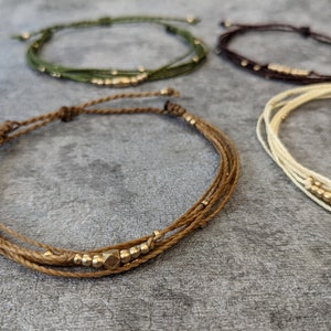 Könnte beinhalten: Vier Armbänder aus dünnen, geflochtenen Schnüren mit goldenen Perlen. Die Armbänder sind in verschiedenen Farben: grün, braun, beige und weiß.