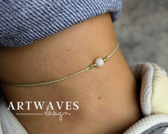 Può includere: Una delicata cavigliera caratterizzata da una singola perlina rotonda bianca affiancata da due piccoli distanziatori metallici dorati su un sottile cordino verde salvia. Il testo ARTWAVES design è stampato in bianco in basso a sinistra.