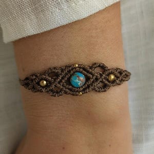 Puede incluir: Un brazalete de macramé marrón con una piedra preciosa turquesa y cuentas doradas. El brazalete está atado en un patrón de nudos.