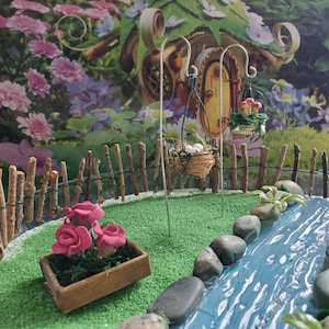 May include: A miniature fairy garden scene with a small wooden fence, a stream, and three bird nests hanging from metal hooks. The nests are made of twigs and have white eggs inside. There are pink flowers in a wooden planter box.