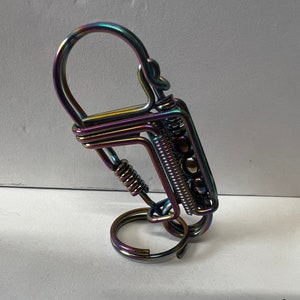 May include: Iridescent metal keychain with a spring and decorative beads. The keychain has a large loop for keys and a smaller loop at the bottom. The metal has a rainbow finish, reflecting colors like purple, green, and gold.