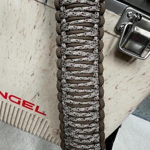 Pode incluir: Close-up de uma pulseira de paracord marrom e branca. A pulseira &eacute; intrincadamente tecida com um padr&atilde;o repetitivo de fios marrons, brancos e salpicados. As bordas s&atilde;o de cor marrom s&oacute;lida. A pulseira est&aacute; em uma superf&iacute;cie clara.