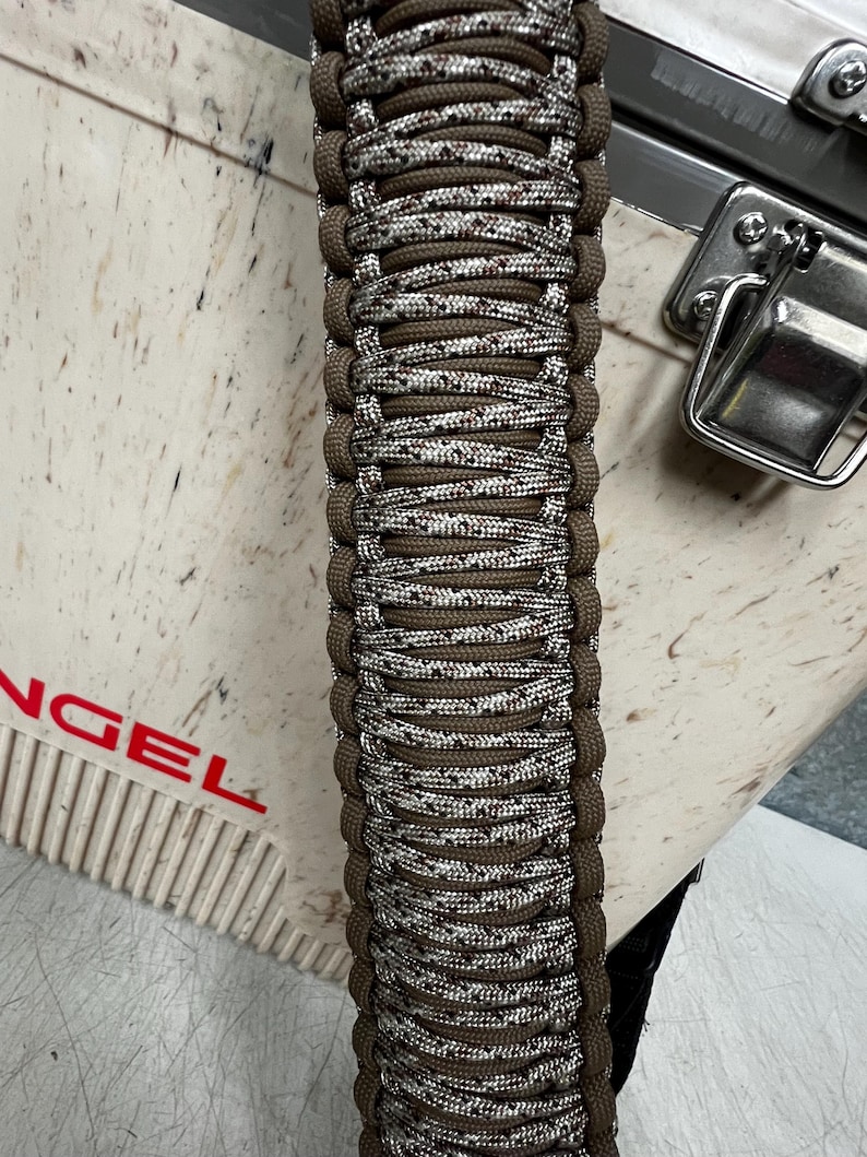 Pode incluir: Close-up de uma pulseira de paracord marrom e branca. A pulseira &eacute; intrincadamente tecida com um padr&atilde;o repetitivo de fios marrons, brancos e salpicados. As bordas s&atilde;o de cor marrom s&oacute;lida. A pulseira est&aacute; em uma superf&iacute;cie clara.