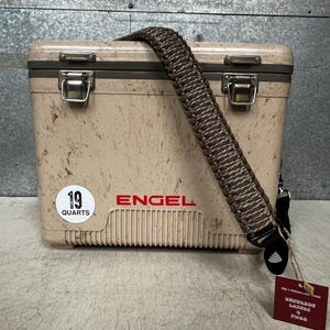 Pode incluir: Uma geleira Engel bege com uma al&ccedil;a tran&ccedil;ada marrom. A geleira tem um padr&atilde;o salpicado e fechos de metal. Um adesivo branco diz "19 QUARTS" e a marca "ENGEL" &eacute; impressa em vermelho.