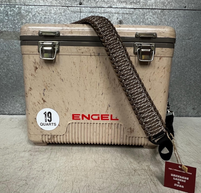 Pode incluir: Uma geleira Engel bege com uma al&ccedil;a tran&ccedil;ada marrom. A geleira tem um padr&atilde;o salpicado e fechos de metal. Um adesivo branco diz "19 QUARTS" e a marca "ENGEL" &eacute; impressa em vermelho.