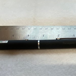 May include: A black laser pointer with a silver band and the text "Renegade Lasers" printed on it. The laser pointer is about 5.5 inches long and is lying on a white surface next to a ruler.