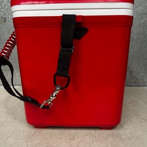 K&ouml;nnte beinhalten: Rote K&uuml;hlbox mit wei&szlig;em Deckel und schwarzem Gurt. Die K&uuml;hlbox hat einen Tragegriff und einen Clip.