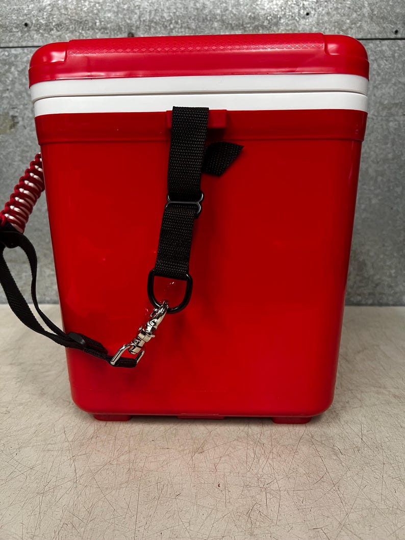 K&ouml;nnte beinhalten: Rote K&uuml;hlbox mit wei&szlig;em Deckel und schwarzem Gurt. Die K&uuml;hlbox hat einen Tragegriff und einen Clip.