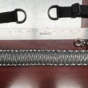 May include: A black and silver tactical rifle sling with two adjustable straps and a metal hook. The sling is made of a braided material and has a woven pattern. The sling is measured against a ruler.
