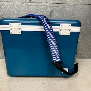 May include: A blue and white cooler with a white and blue braided strap. The cooler has a white stripe around the top and bottom.