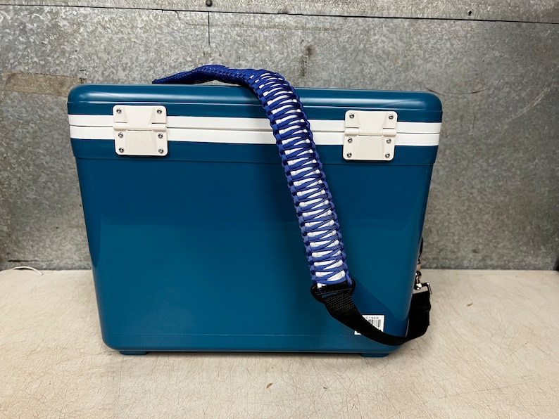 May include: A blue and white cooler with a white and blue braided strap. The cooler has a white stripe around the top and bottom.