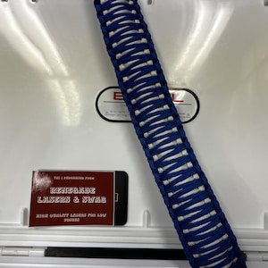 May include: A blue and white paracord handle for a cooler. The handle is made of braided paracord and is about 12 inches long. The handle is lying on top of a white cooler. A business card for "Renegade Lasers & Swag" is visible in the lower left corner of the image.