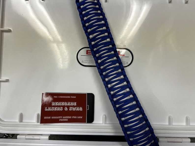 May include: A blue and white paracord handle for a cooler. The handle is made of braided paracord and is about 12 inches long. The handle is lying on top of a white cooler. A business card for "Renegade Lasers & Swag" is visible in the lower left corner of the image.