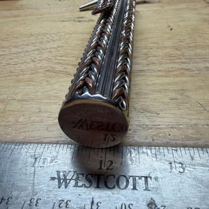 May include: A handcrafted metal object with a braided design in silver and copper tones. The object has a cylindrical shape with a circular end that reads "MEXICO". A metal ruler with inch measurements is in the foreground.