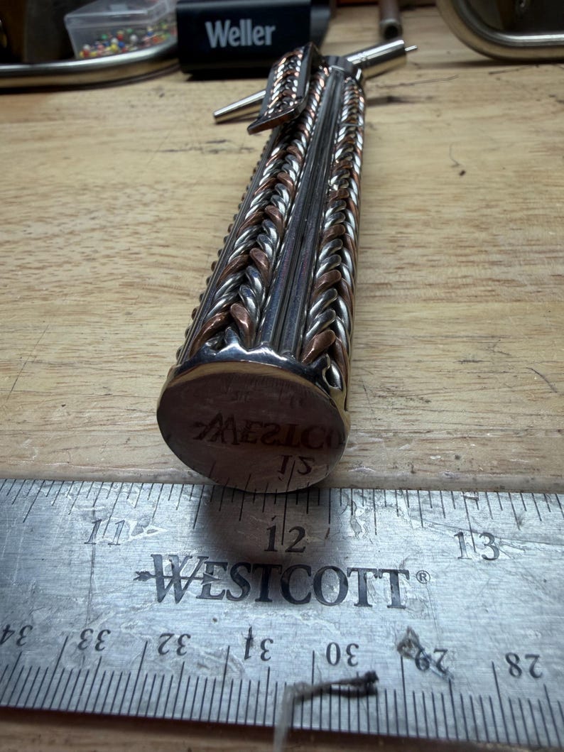 May include: A handcrafted metal object with a braided design in silver and copper tones. The object has a cylindrical shape with a circular end that reads "MEXICO". A metal ruler with inch measurements is in the foreground.