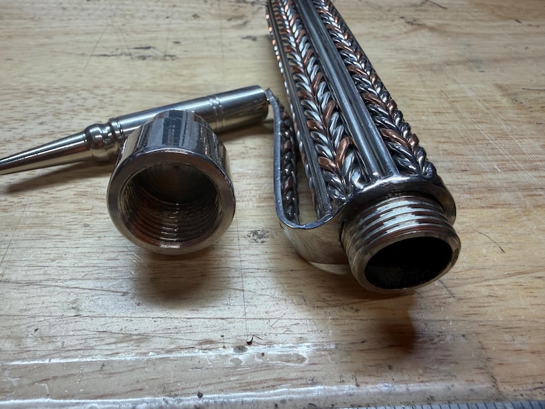 May include: A disassembled metal object with a threaded end, a cylindrical component, and a braided design. The object is silver and copper-colored, and appears to be a part of a larger mechanism or tool.