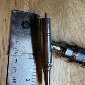 May include: A shiny, silver metal tap handle, approximately 3 inches long, with a pointed end and a cylindrical body. The handle is next to a metal ruler on a wooden surface. The tap handle is a component of a beer tap system.
