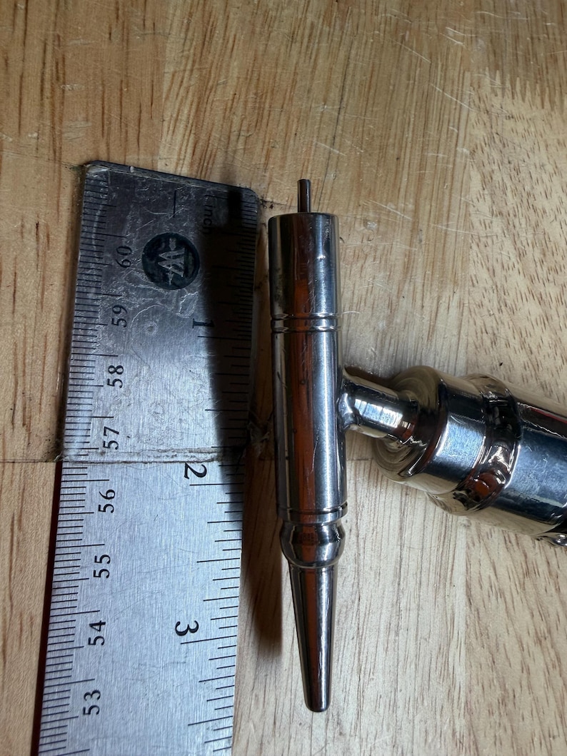 May include: A shiny, silver metal tap handle, approximately 3 inches long, with a pointed end and a cylindrical body. The handle is next to a metal ruler on a wooden surface. The tap handle is a component of a beer tap system.