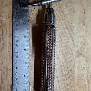 May include: A metal corkscrew with a decorative handle. The handle is made of interwoven copper and silver-colored metal. A metal ruler is next to the corkscrew, showing a length of approximately 8 inches.