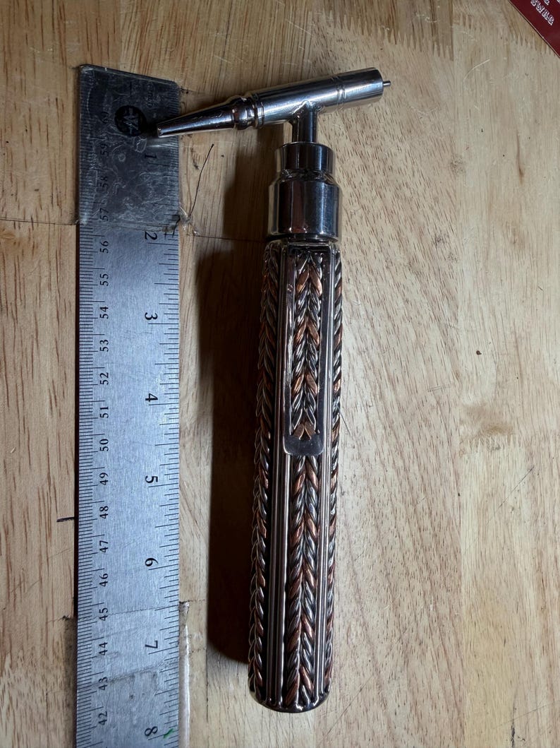 May include: A metal corkscrew with a decorative handle. The handle is made of interwoven copper and silver-colored metal. A metal ruler is next to the corkscrew, showing a length of approximately 8 inches.