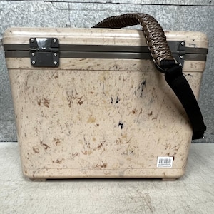 Pode incluir: Uma geleira bege com um padr&atilde;o salpicado, com uma al&ccedil;a tran&ccedil;ada marrom e uma al&ccedil;a de ombro preta. A geleira tem formato retangular com tampa articulada e fechos de metal. Projetada para transportar itens.