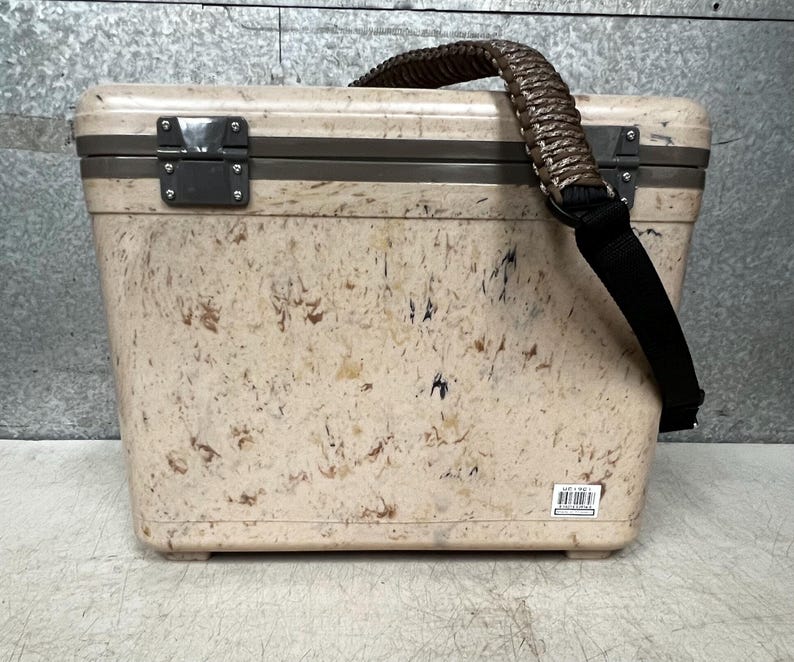 Pode incluir: Uma geleira bege com um padr&atilde;o salpicado, com uma al&ccedil;a tran&ccedil;ada marrom e uma al&ccedil;a de ombro preta. A geleira tem formato retangular com tampa articulada e fechos de metal. Projetada para transportar itens.