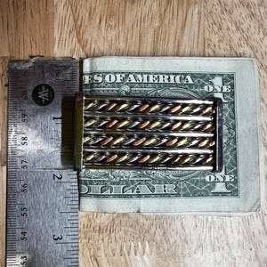 May include: A rectangular money clip with a silver frame and a series of twisted metal bars in silver, gold, and copper tones. The clip is holding a one-dollar bill. A metal ruler is next to the money clip.
