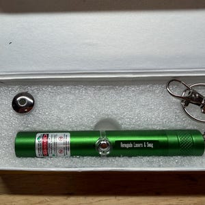 May include: A green laser pointer with the text "Renegade Lasers & Swag" in a white box. The box contains a silver-colored metal cap and a silver keychain. The laser pointer has a warning label.