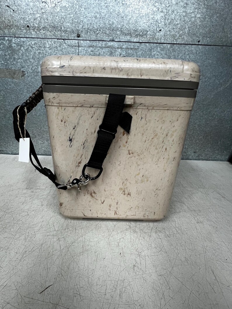 Pode incluir: Um cooler bege com um padr&atilde;o salpicado, uma al&ccedil;a de ombro preta e uma al&ccedil;a preta. O cooler tem formato retangular com uma moldura cinza ao redor da tampa. Uma etiqueta branca pende da al&ccedil;a.