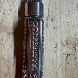May include: Close-up of a metal bottle opener with a braided handle. The handle is a mix of silver and copper-colored metal strands. The top portion of the opener is a shiny silver color.