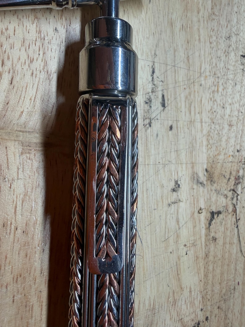 May include: Close-up of a metal bottle opener with a braided handle. The handle is a mix of silver and copper-colored metal strands. The top portion of the opener is a shiny silver color.