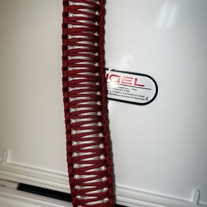 K&ouml;nnte beinhalten: Ein roter und wei&szlig;er Paracord-Griff f&uuml;r eine K&uuml;hlbox. Der Griff ist aus geflochtenem Paracord gefertigt und an einer wei&szlig;en K&uuml;hlbox befestigt. Die K&uuml;hlbox tr&auml;gt ein Etikett mit der Aufschrift "Engel" und ein Warnsymbol.