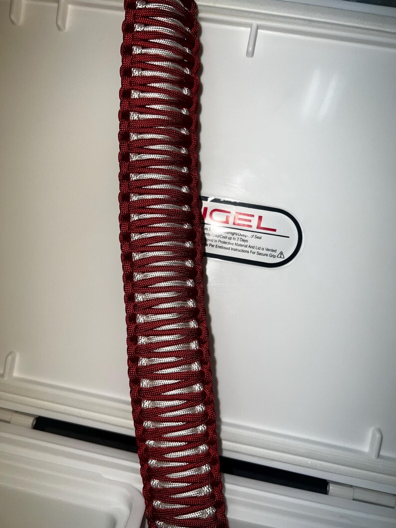 K&ouml;nnte beinhalten: Ein roter und wei&szlig;er Paracord-Griff f&uuml;r eine K&uuml;hlbox. Der Griff ist aus geflochtenem Paracord gefertigt und an einer wei&szlig;en K&uuml;hlbox befestigt. Die K&uuml;hlbox tr&auml;gt ein Etikett mit der Aufschrift "Engel" und ein Warnsymbol.